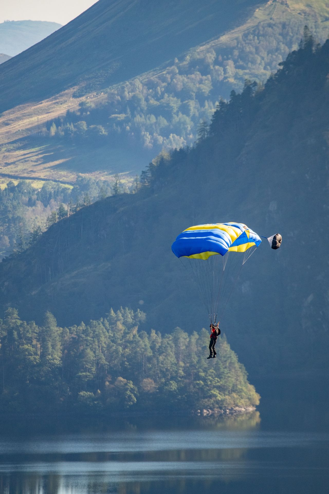 A base jumper descending with a blue and yellow parachute over a calm reservoir, with forested hills and rugged mountains in the background