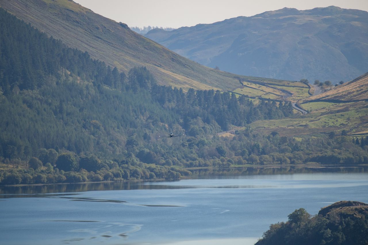 Scenic view of Thirlmere Reservoir surrounded by forested hills and rolling countryside, with a small Texan T1 aeroplane flying low over the water, set against a backdrop of distant mountains.