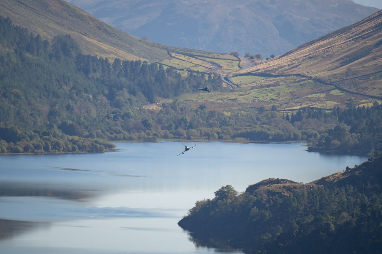 Two Eurofighter Typhoon jets flying low over a serene reservoir, surrounded by forested hills and a winding countryside road, with distant mountains in the background.