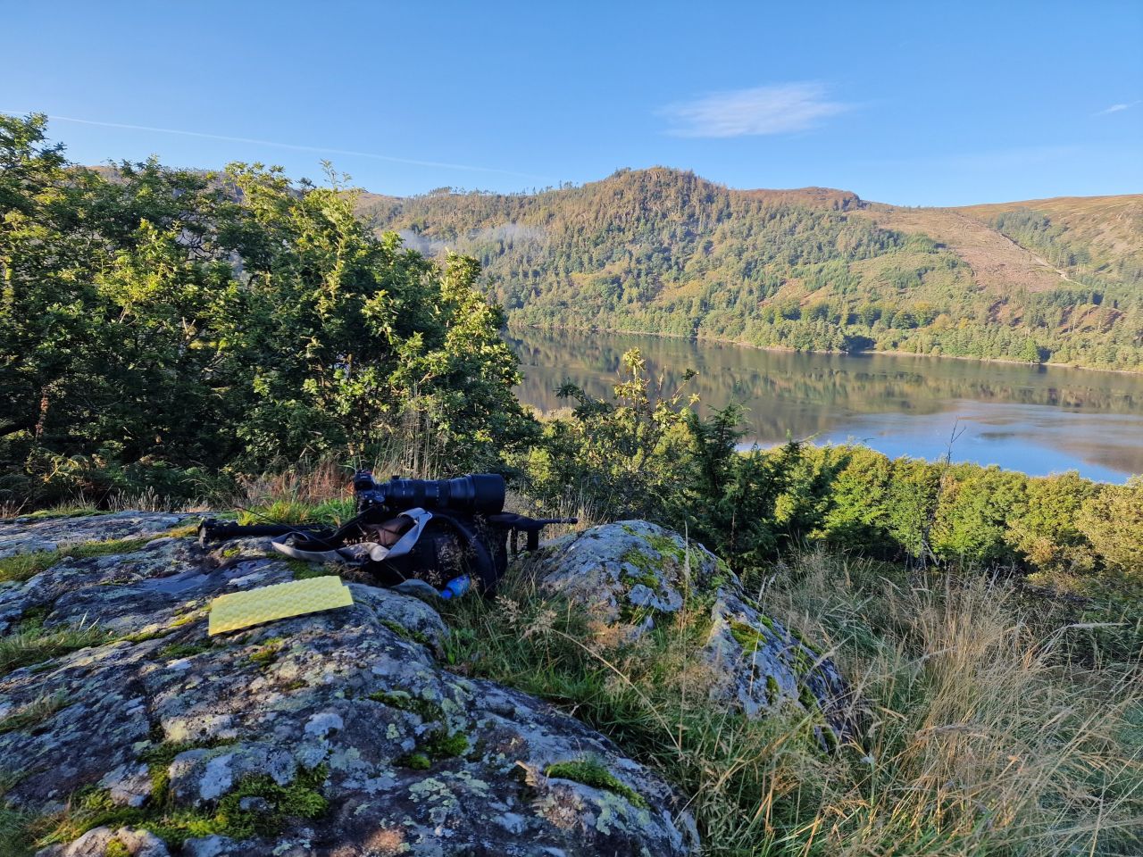 Camera equipment, including a large telephoto lens and backpack, resting on a moss-covered rock with a scenic view of a calm reservoir surrounded by hills and forests in the background.