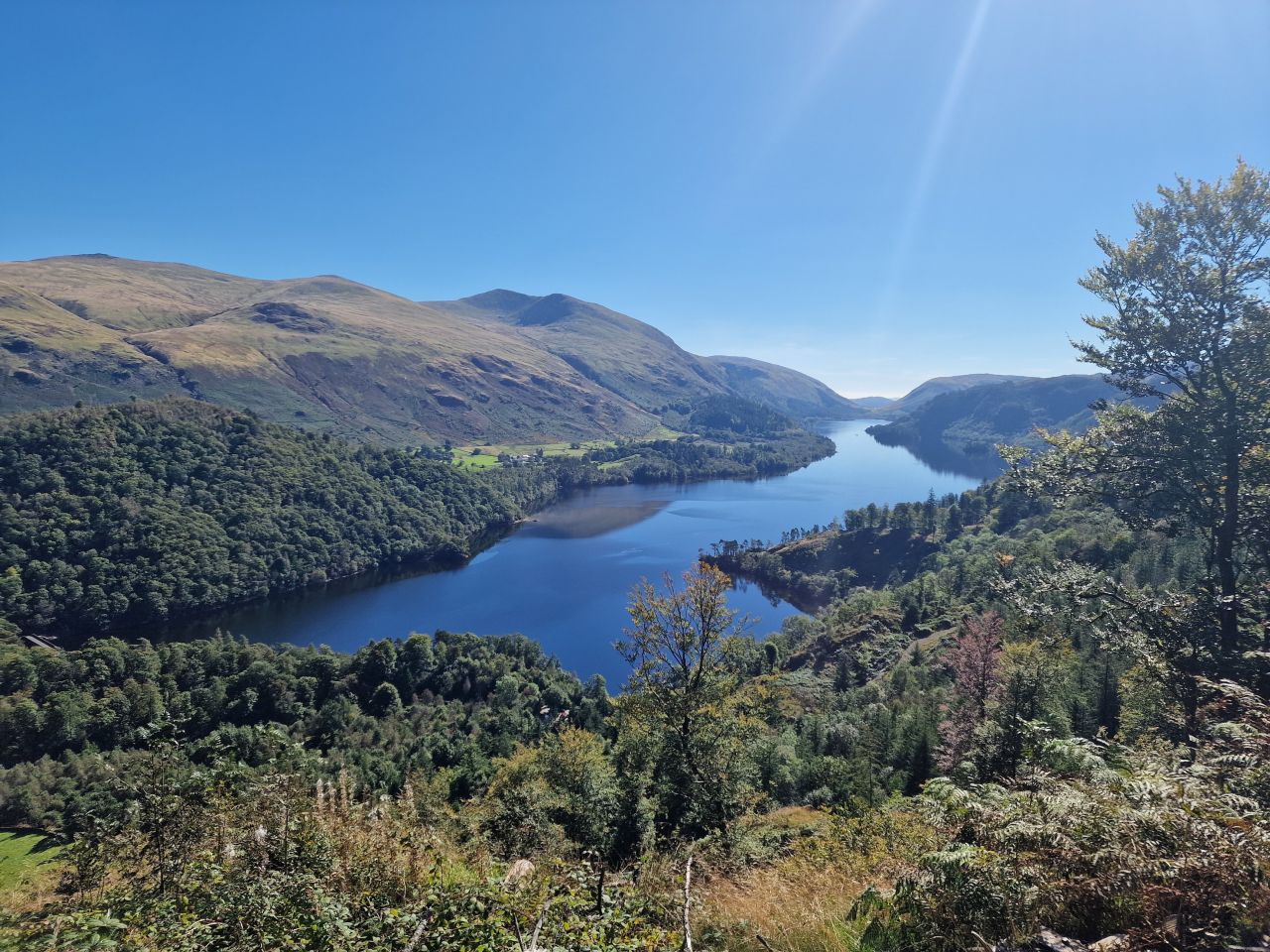 Panoramic view of Thirlmere Reservoir, surrounded by lush green hills and mountains under a bright blue sky, with sunlight casting a gentle glow over the landscape.