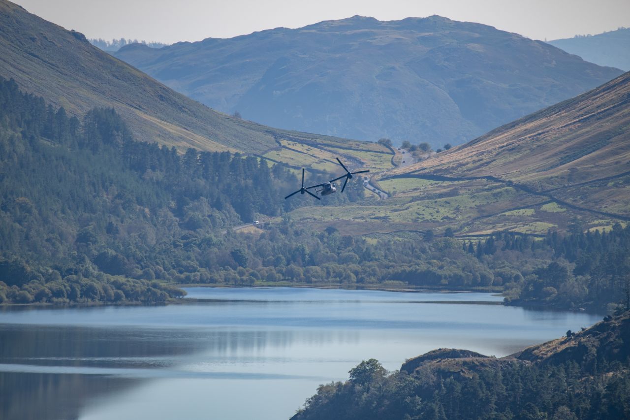 V-22 Osprey flying low over Thirlmere Reservoir, surrounded by rolling hills and forests. A winding road runs through the lush, mountainous landscape, with the calm waters of the reservoir reflecting the natural scenery.