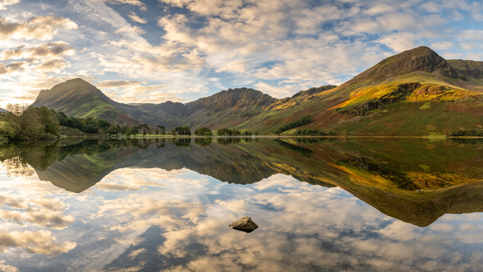 This image showcases a tranquil lake with perfectly still water, reflecting the surrounding mountains and sky. The landscape features towering, green-covered mountains under a partly cloudy sky, creating a symmetrical scene with the reflection. A single rock is visible in the foreground in the clear water, adding to the sense of calm and balance in the composition. Trees line the far edge of the lake, contributing to the serene natural setting.