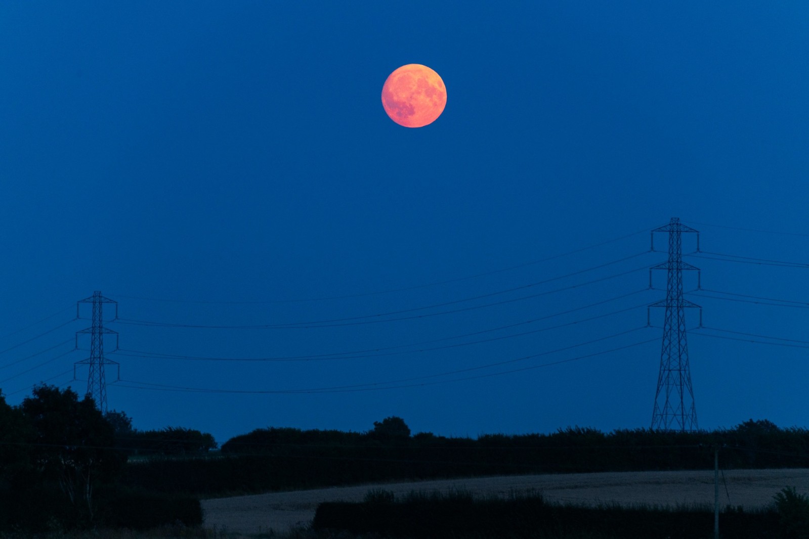 A vibrant red supermoon rises against a deep blue evening sky, framed by two power lines and tall pylons in a rural landscape.