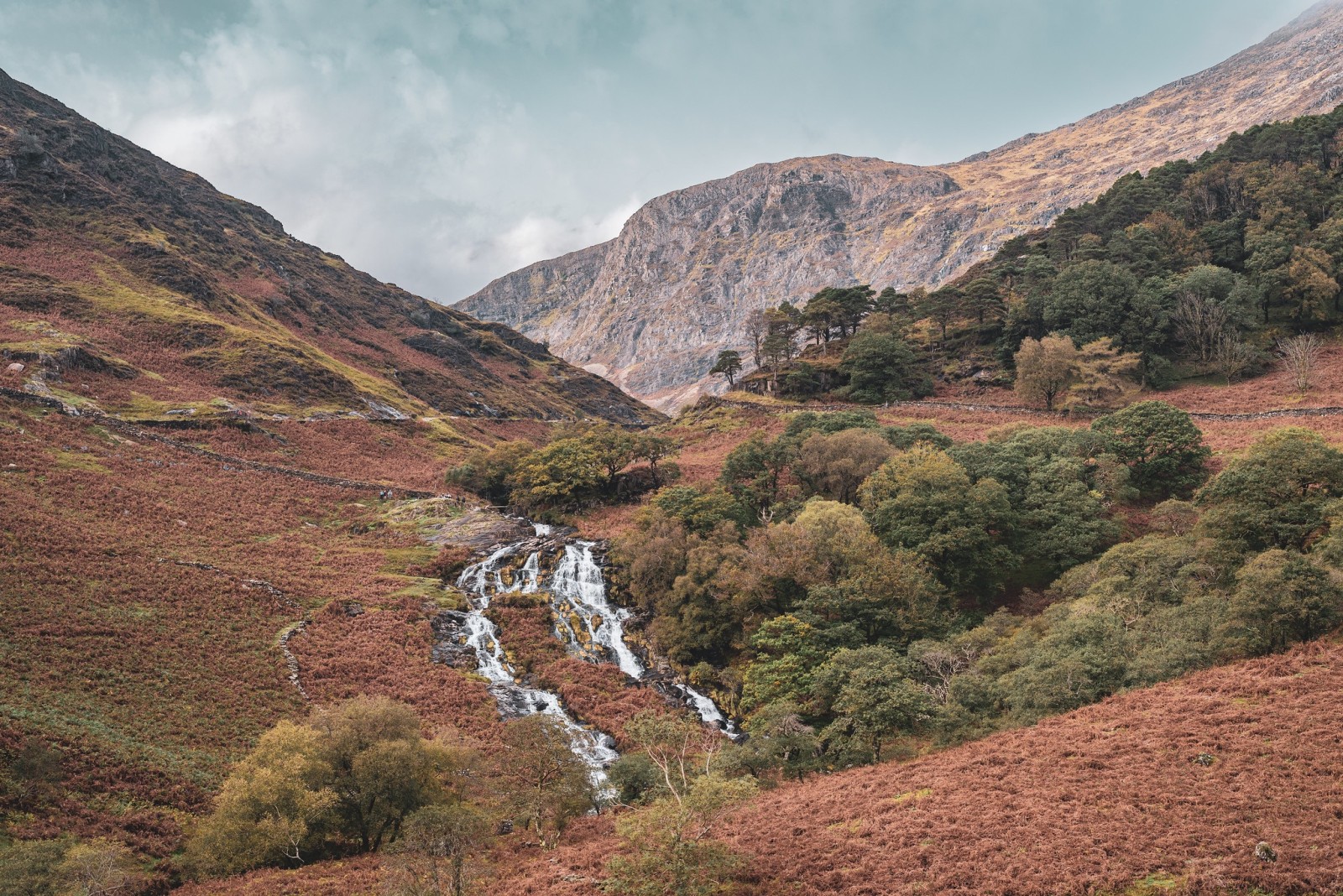 A scenic view of a valley along the path to Snowdon in Wales, featuring a small waterfall flowing down the hillside. The landscape is dotted with autumn-colored foliage, green trees, and rocky hills, with a backdrop of towering mountains under a cloudy sky.