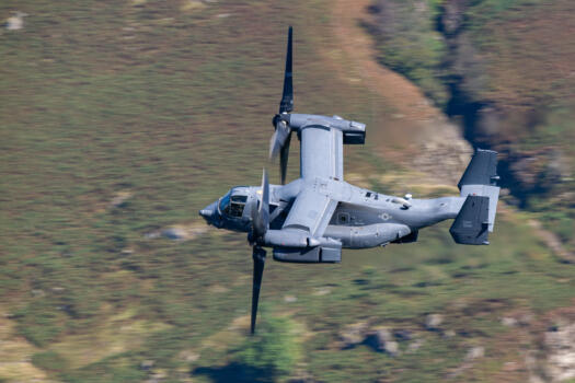 Close-up of a V-22 Osprey in flight, featuring its distinct tilt-rotor design and grey military livery, flying low over a green, hilly terrain