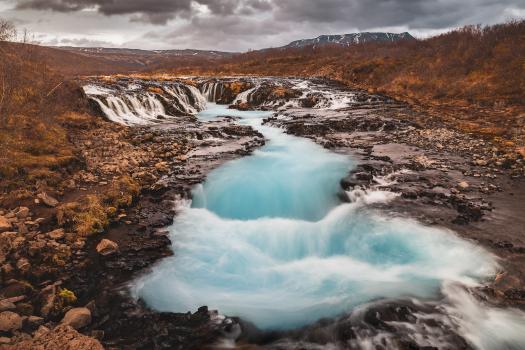 A stunning view of Bruarfoss Waterfall in Iceland, featuring bright turquoise water cascading over dark, rocky terrain. The water flows through a narrow channel, creating multiple small falls before reaching a pool below. The surrounding landscape is rugged with autumnal tones and distant mountains under a cloudy sky.