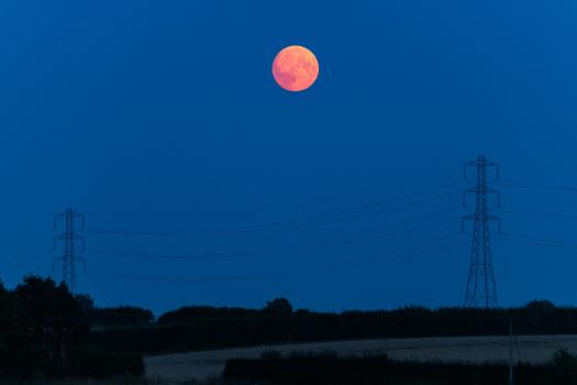 A vibrant red supermoon rises against a deep blue evening sky, framed by two power lines and tall pylons in a rural landscape.