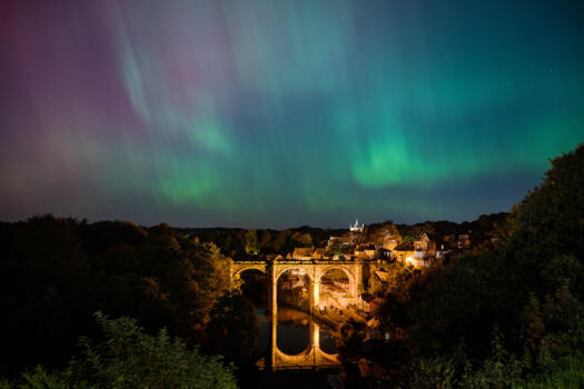 This image shows a beautiful scene of the Northern Lights over a train viaduct in Knaresborough, a picturesque town known for its historic architecture and scenic beauty. The aurora lights in shades of green and purple blend with the night sky, creating a stunning contrast with the warmly lit viaduct and surrounding landscape. The viaduct's reflection in the river adds to the beauty of the photograph.