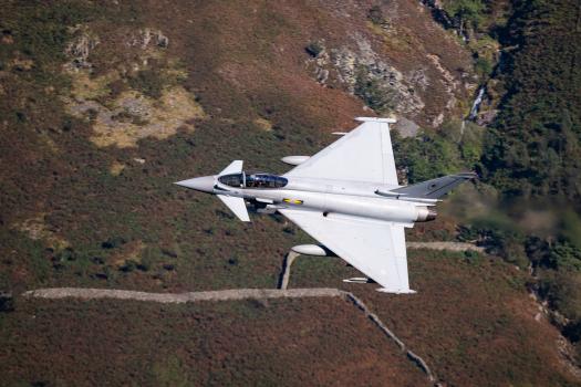 Close-up of a Eurofighter Typhoon jet flying low over a rocky and grassy hillside, showcasing its sleek design and grey military livery.